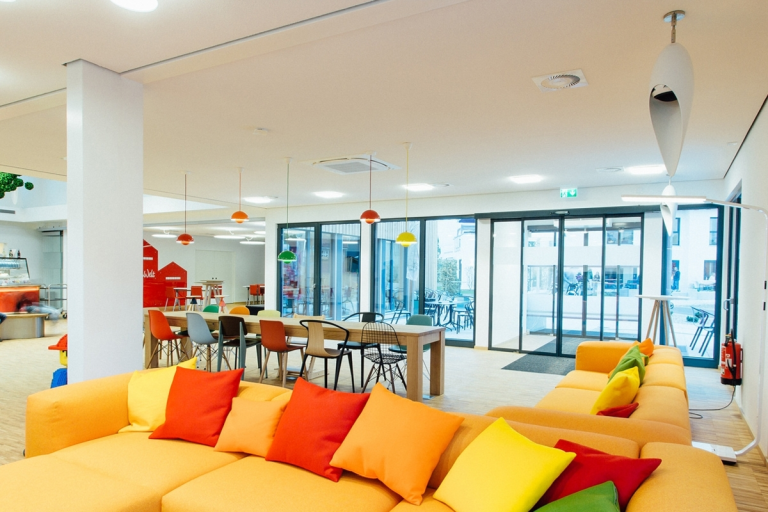 Designlautsprecher Hellblauer, moderner Raum mit bunten Sofakissen und einem Essbereich. Große Fenster sorgen für Licht.