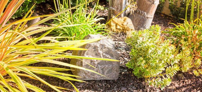 Steinlautsprecher Stein umgeben von bunten Pflanzen und Gras in einem Garten.
