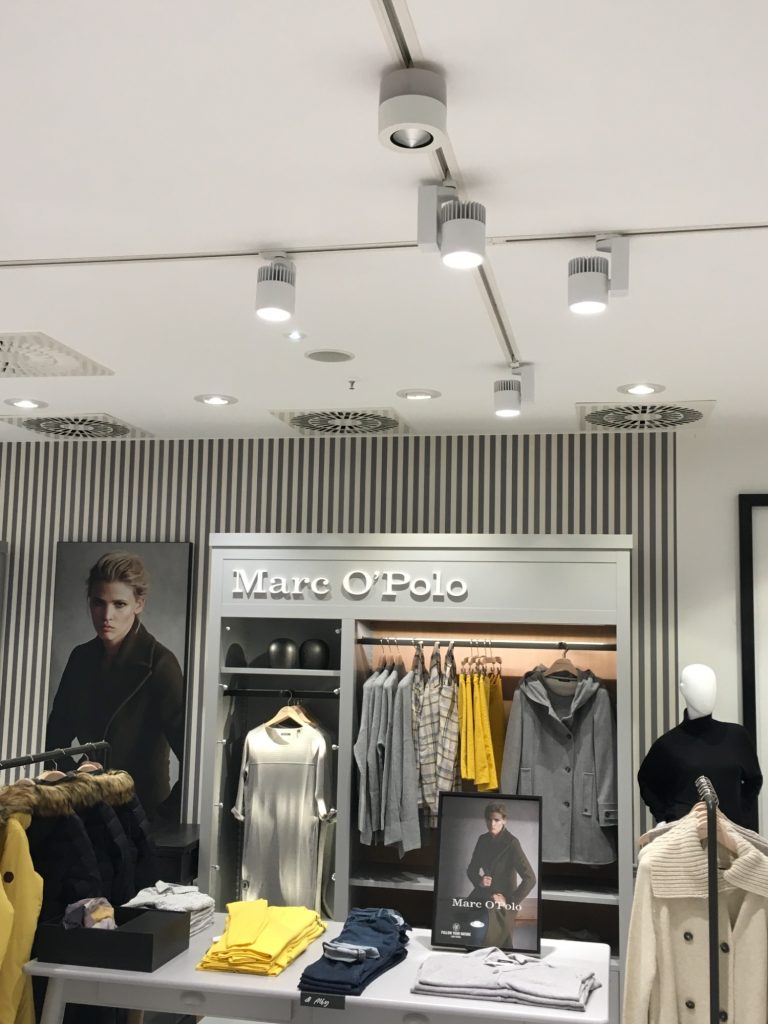 Stromschienenlautsprecher Kleidungsgeschäft mit Hängemöglichkeiten und beleuchtetem Ältere-Model-Poster.