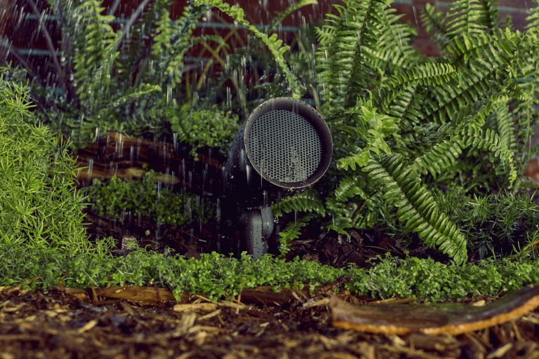 Erdspießlautsprecher Ein Bodenstrahler steht zwischen grünen Pflanzen im Regen.