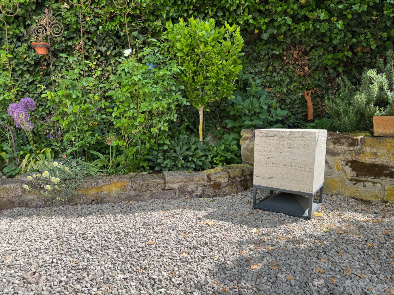 Bodenlautsprecher Ein Gartenbereich mit einer Hecke, einer kleinen Pflanze und einem Quarzkiesboden.